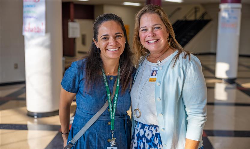 Two educators, one wearing dark blue and the other light blue, smile while standing in a school hallway